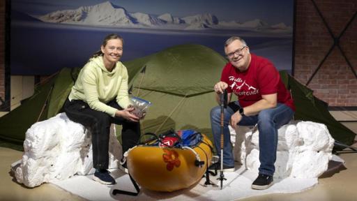 "1, 2 oder 3 - Abenteuerinnen": Birgit Lutz und Elton sitzen im Studio vor einem Zelt auf nachgebauten Eisblöcken. Zwischen ihnen steht ein Kanu.