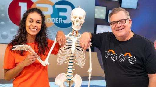 Studiogast Dr. Florence Randrianarisoa (l.) steht neben einem Skelett und hält den rechten Arm. Elton (l.) stützt sich auf die Schulter des Skeletts.