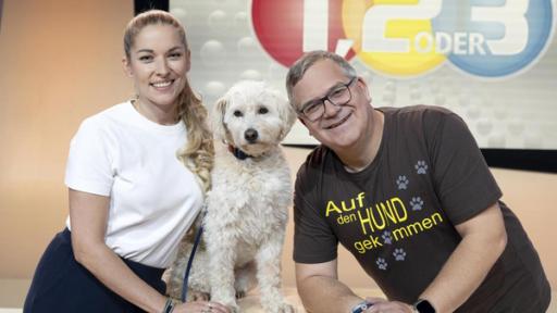 Conny Sporrer und Elton stehen links und rechts neben einem Podest. Auf dem Podest sitzt Connys niedlicher Hund. 