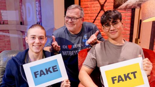 Vico und Dominik sitzen auf Sesseln und lächeln. Vico hält ein Schild, auf dem "Fake" steht, und Dominik ein Schild, auf dem "Fakt" steht. Elton kniet dahinter und lächelt.