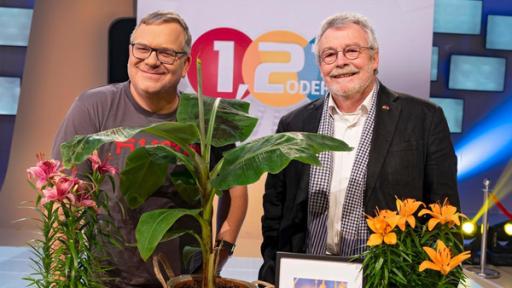 Elton und sein Gast Prof. Dr. Hans Leisen stehen neben einem Tisch mit Gegenständen, die typisch für Kambodscha sind. Auf dem Tisch stehen zum Beispiel Pflanzen, Bananen, Getränkedosen und die Nationalflagge Kambodschas. Elton hält eine Mango in der Hand.