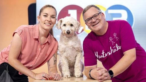 Conny Sporrer (l.) stützt sich auf ein Pult und lächelt. Rechts Daneben steht Elton auf das gleiche Pult gestützt. In der Mitte sitzt ein Hund.