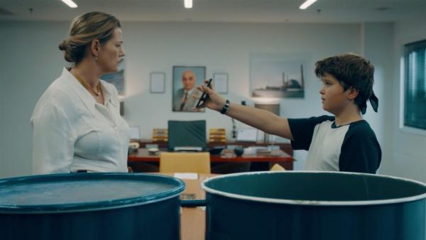 Sammy (Arthur Hermans) hält Patricia (Emilie De Roo) sein Handy vor die Nase. Beide stehen in einem Büro hinter zwei blauen Metallfässern.