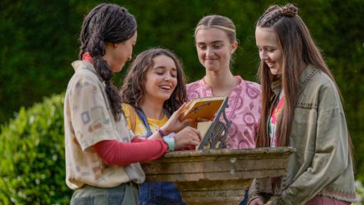 Audrey, Addie, Emily und Jenna stehen draußen im Freien um einen runden Tisch aus Stein herum. Sie schauen alle lachend auf das Notizbuch, das Addie in ihren Händen hält.