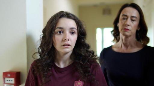 Böse ist, wer Böses tut - Folge 8 | Rechte: Kind Of Productions Limited MMXXIII In einem weiß-gestrichenem Schulkorridor steht Addie mit fassungslosem und schockiertem Blick. Etwas versetzt hinter ihr steht die Lehrerin Miss Murphy. Sie schaut mit strengem Blick auf Addie herab.