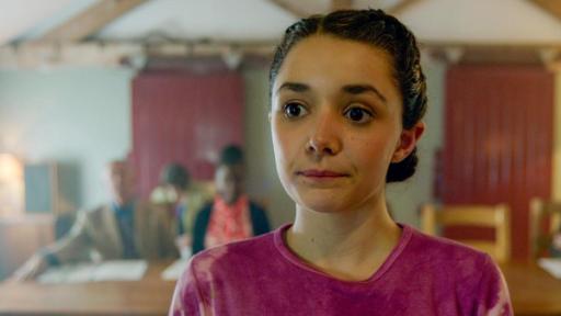 Das Denkmal - Folge 2 | Rechte: Kind Of Productions Limited MMXXIII Addie steht im Vordergrund und schaut leicht an der Kamera vorbei. Im Hintergrund sitzen 3 Erwachsene an einem langen Holztisch mit Papieren vor sich liegend.