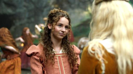 Die Befreiung - Folge 10 | Rechte: Kind Of Productions Limited MMXXIII Im 16. Jahrhundert draußen stehen sich die Schwestern Maggie (Hattie Gotobed) und Elinor (Ella Maisy Purvis) in ihren edlen Kleidern gegenüber. Maggie schaut ernst aber liebevoll mit leicht geneigtem Kopf zu Elinor, die nur von hinten zu sehen ist. Der Hintergrund ist unscharf, doch es sind tanzende Kinder in rostroten Kleidern zu erkennen.