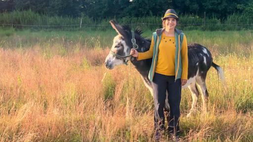 Anna steht lächelnd auf einer Wiese und hält einen Esel am Halfter