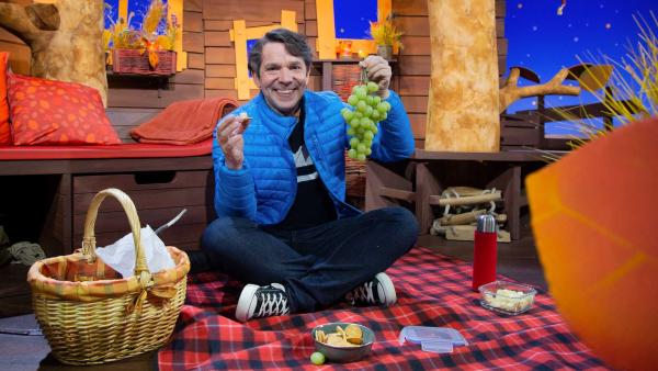 Juri sitzt auf einer Decke und hat ein Picknick ausgebreitet. Er hat Käse und Trauben in der Hand.