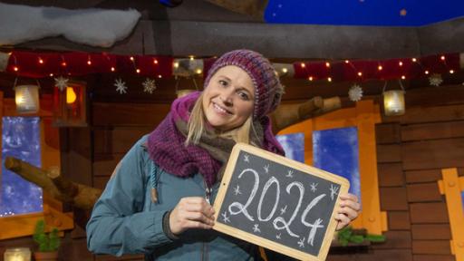 Singa steht im Baumhaus. Sie hält eine Kreidetafel in der Hand, auf welcher das Jahr 2024 steht.