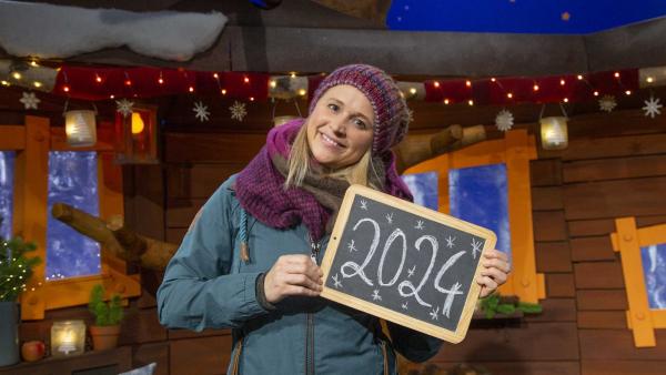 Singa steht im Baumhaus. Sie hält eine Kreidetafel in der Hand, auf welcher das Jahr 2024 steht.