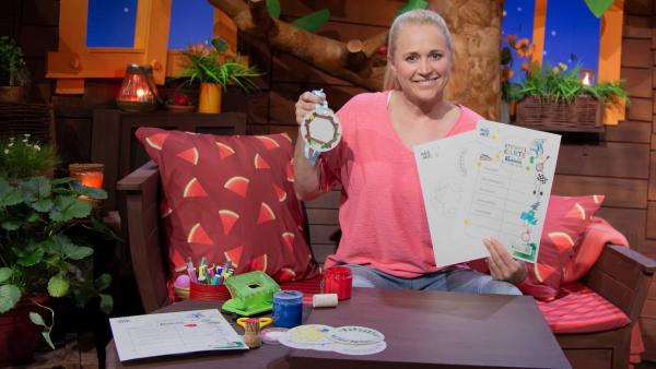 Singa sitzt auf dem Sofa. Sie hält die MACH MiT!-Bastelbögen und eine Medaille in der Hand.