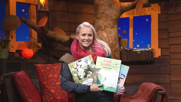 Singa sitzt auf dem Sofa im Baumhaus und hält drei Kinderbücher in der Hand.