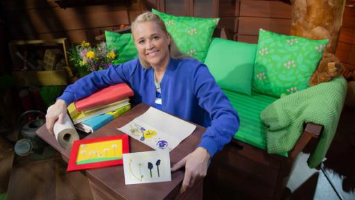 Rechte: KiKA Singa hat verschiedene Bücher, eine Küchenrolle und bunte Blüten vor sich auf dem Tisch liegen.