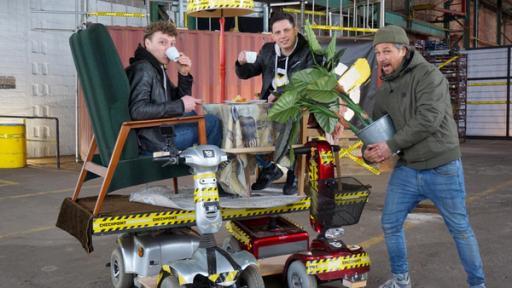 Im Bild befinden sich zwei Senioren-Scooter, auf denen längs eine Platte angebracht ist. Darauf stehen zwei Sessel, in denen zwei Checkpoint-Tester gemütlich Tee trinken. Tommy Scheel steht rechts im Bild und hält eine Zimmerpflanze in der Hand. 