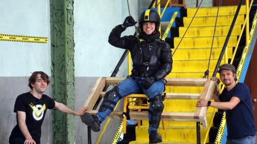 Checkpoint: Tommy und das Testteam stehen an einer Treppe. Ein Checkpoint-Mitglied schwebt in einem Gurt über der Treppe. 