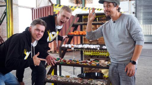 Checkpoint: Tommy Scheel steht rechts im Bild und hält eine Murmel in der Hand. Jeppe und Mark knien vor einer Treppe, auf der verschiedene Gegenstände liegen.