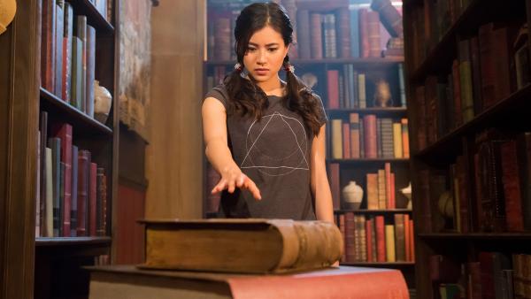 Kyra (Kimie Tsukakoshi) hat in der Bibliothek der Magie einen Stapel alter Bücher entdeckt. Vom obersten Buch wird sie magisch angezogen. | Rechte: ZDF/Jonathan M. Shiff Productions/Screen Queensland