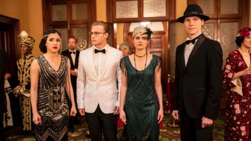 Ein verzweifelter Plan | Rechte: ©2020 Jonathan M. Shiff Productions Kyra (Kimie Tsukakoshi), Peter (Jamie Carter), Imogen (Elizabeth Cullen) und Darra (Julian Cullen) stehen in festlicher Kleidung in der Mode von 1919 nebeneinander in einer Empfangshalle zwischen anderen Gästen. Sie schauen sich angespannt um.