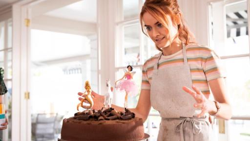 Eine ganz besondere Torte | Rechte: ©2020 Jonathan M. Shiff Productions Mathilda (Arnijka Larcombe-Weate) steht vor einer Schokoladentorte, die mit drei Figuren verziert ist, einer Fee, einer Meerjungfrau und einem Einhorn. Mathilda hat eine Küchenschürze umgebunden und schaut erfreut auf die Torte vor ihr. Hinter ihr sieht man eine helle Fensterfront.