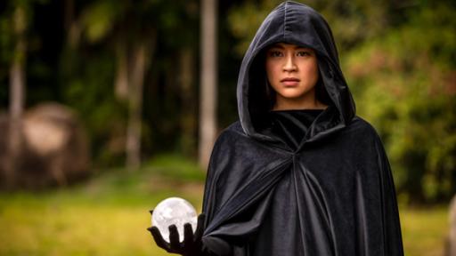 Ungewohnte Zaubertricks | Rechte: ©2020 Jonathan M. Shiff Productions Kyra (Kimie Tsukakoshi) steht, in einen schwarzen Kapuzenumhang gehüllt, in einem Wald. Sie hält in der rechten Hand eine weiße Kugel und blickt ernst nach vorne.