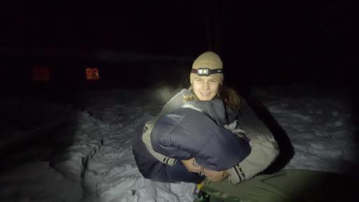 Emilia ist warm bekleidet. Sie sitzt in einem Schlafsack im Schnee. Ihre Kopflampe leuchtet, die Umgebung ist nachtdunkel.