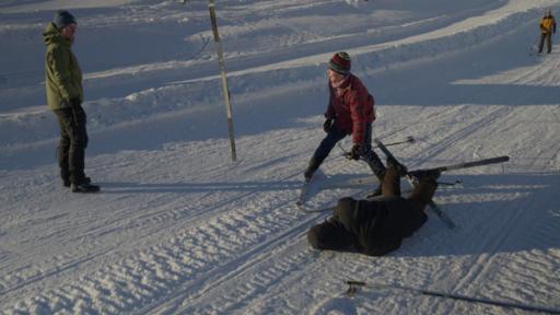 Drei Personen befinden sich auf einer Skipiste. Zwei von ihnen haben Ski an ihren Füßen. Die rechte Person ist hingefallen.