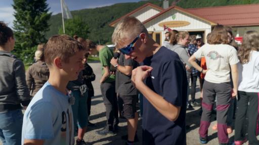 Im Vordergrund unterhalten sich zwei Jungen. Im Hintergrund stehen mehrere Personen. 