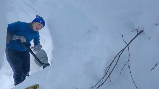 Kaja steht in Winterklamotten in einer Schneehöhle. In der Hand hält sie eine Schaufel.