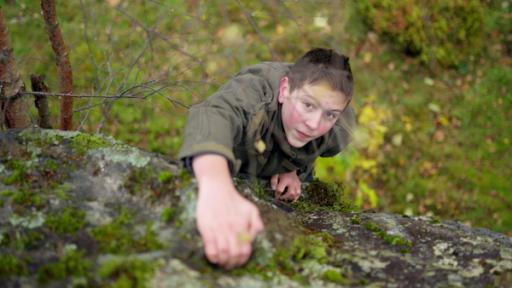 Ben steht auf Waldboden und blickt aufwärts. Mit der Hand sucht er Halt an einem Felsen.