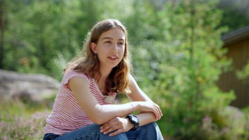 Emma sitzt in der Landschaft und schaut in die Ferne. Verschwommen sind im Hintergrund Bäume und eine Hütte zu sehen.