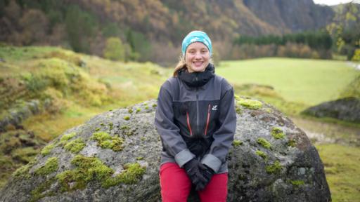 Kaja lehnt an einem Fels und schaut lächelnd in die Kamera. Im Hintergrund ist eine Wiesen- und Berglandschaft zu sehen.