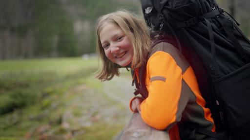 Leyla lehnt über einem Geländer im Wald. Sie schaut lächelnd in die Kamera. Auf dem Rücken trägt sie einen großen Rucksack.