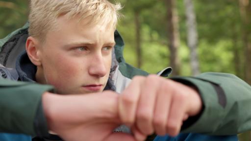 Maximilian sitzt nah an der Kamera im Wald und schaut in die Ferne. Hinter ihm sind verschwommene Bäume zu sehen.