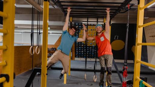 "Das Wunder DU - Ganz schön stark": Christoph (l.) und Angelino (r.) hängen an einem Arm unter dem Dach einer Kletterhalle. Mit der anderen Hand klatschen sie sich ab. beide lachen.