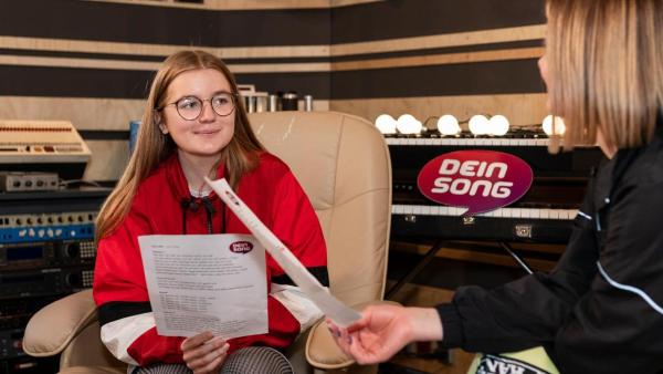 "Dein Song - Gemeinsam kreativ sein - Folge 14": Fine (links) sitzt auf einem Sessel im Tonstudio und hört lächelnd ihrer Patin Mathea zu, die rechts von ihr sitzt. Beide halten Textblätter in der Hand. | Rechte: ZDF
