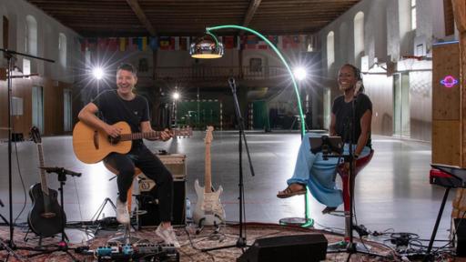 Die vierköpfige "Dein Song"-Band sitzt lachend in einer großen Halle. Sie sind umgeben von ihren Instrumenten.