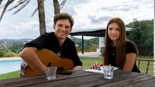 KAMRAD (links) und Malaika (rechts) sitzen an einem Tisch und gucken direkt in die Kamera. KAMRAD hat eine Gitarre auf dem Schoß liegen, vor Malaika liegen mehrere Notizblätter.