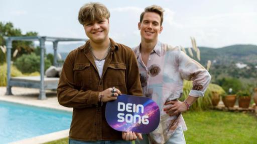 Felix und Conny stehen nebeneinander. Felix hält ein Dein Song-Schild in den Händen, CONNY hat seinen rechten Arm in die Hüften gestemmt. Beide lächeln.