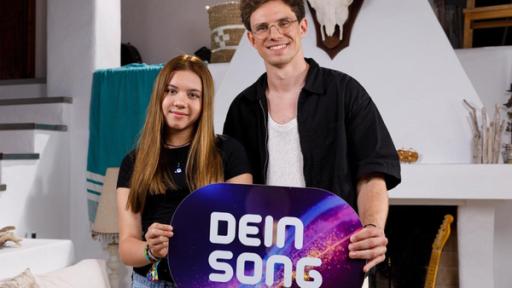 Maleika und Kamrad stehen nebeinander. Mit ihren Händen halten sie auf Bauchhöhe ein Dein Song-Schild. Beide lächeln. 