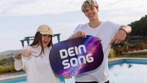 Luna und Constantin stehen nebeneinander und halten ein Dein Song-Schild in den Händen, auf das sie mit ihren Zeigefingern deuten. Beide lächeln.