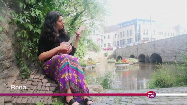 Rona sitzt mit einer Ukulele in der Hand an einem Fluss. | Rechte: KiKA