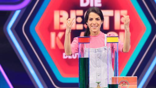 Im Studio. Clarissa lächelnd vor dem Logo "Die beste Klasse Deutschlands". Vor ihr auf einem Tisch zwei große Reagenzgläser, die mit grüner Flüssigkeit gefüllt sind.    