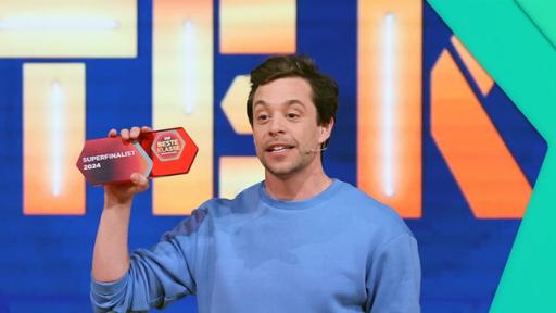 Mittig steht Tobi. Er hält in seiner Hand ein kleines Schild aus Plastik mit der Aufschrift Superfinalist 2024 und dem Logo von Die beste Klasse Deutschlands. Im Hintergrund große Buchstaben.