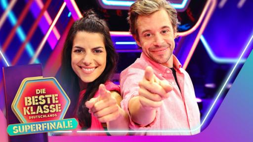 Clarissa und Tobi stehen lächelnd eng nebeneinander und zeigen mit dem Finger nach vorne. Damit machen sie auf das Superfinale der beliebten Quizshow aufmerksam.