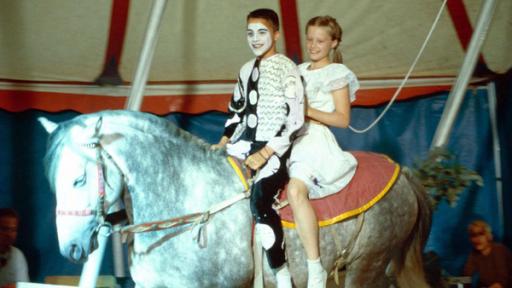 Rafael als Harlekin und Natascha reiten im Zirkuszelt auf einem weißen Pony.