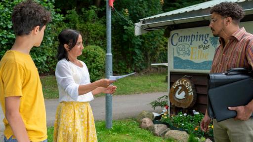 Tarun und seine Mutter Jaswinder stehen vor dem Eingang zum Campingplatz und überreichen einem Mann einen Zettel.