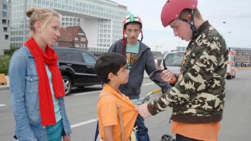 Jessi (Martha Fries) schaut zu einem Jungen mit BMX-Helm (David Riske), der eine Sonnenbrille in der Hand hält und Luis (Emilio Sanmarino, Mitte) am T-Shirt packt. Ein weiter Junge mit Helm schaut zu (Benjamin Falk, hinten)