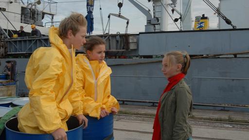 Jessi (Martha Fries, rechts) steht vor Max (Bruno Alexander) und Nina (Carolin Garnier), die mit Schutzanzügen in einer Tonne stehen, am Kai.