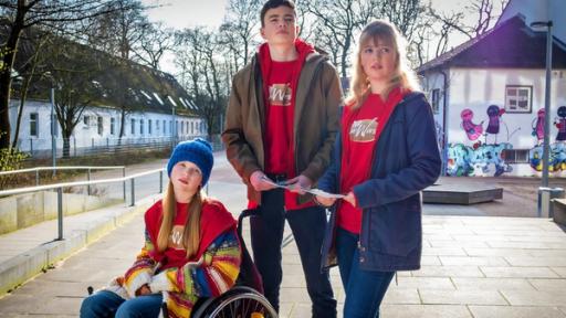 Pippa, Fritz und Paula tragen rote Pullover und verteilen Flyer auf dem Schulhof.